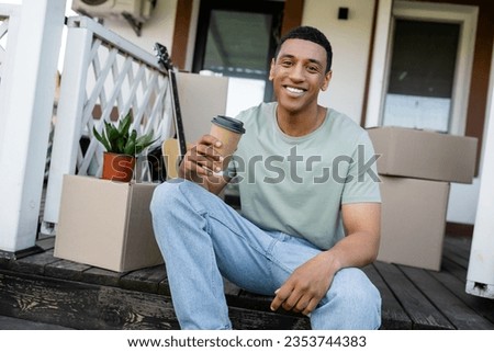 cheerful african american man holding coffee to go near carton boxes and new house outdoors
