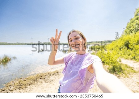 Happy young millenial woman with long brown hair taking a selfie portrait hiking along a forest lake trail, Pov of a smiling brown guy using smart phone mobile to take a picture of herself on holiday