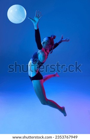 Motivated young woman, volleyball player during game, hitting ball in jump against blue studio background in neon light. Concept of professional sport, competition, health, hobby, action, ad