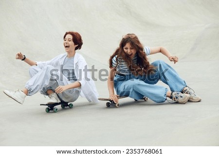 Laughing, cheerful, happy teen girls, friends in casual clothes having fun on skate park, sitting and riding skateboard. Concept of youth culture, sport, dynamic, extreme, hobby, action and motion