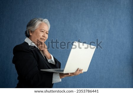 Portrait of intelligent senior businesswoman CEO manager holding in hands laptop creating a presentation on isolated blue background. Old Woman thinking after getting financial statements reports.