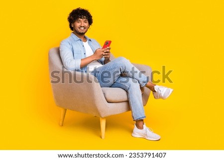 Full size photo of nice positive man dressed denim shirt stylish jeans sit in armchair hold smartphone isolated on yellow color background