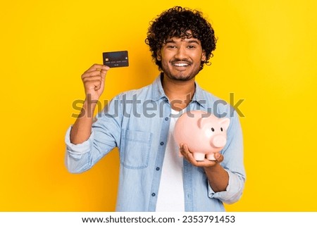 Photo of positive nice young man beaming smile arms hold money bank pig debit plastic card isolated on yellow color background