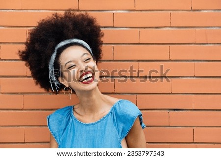 Positive young African American female with Afro hair headband red lips smiling and looking away while standing alone against brown brick wall building in sunny daylight Royalty-Free Stock Photo #2353797543