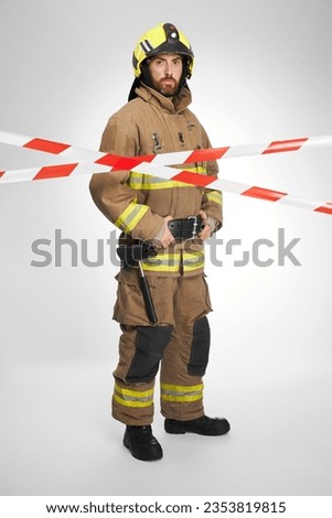 Serious firefighter in full gear standing behind red and white signal tape. Front view of bearded rescuer looking at camera from closed by caution tape area, on white background. Workplace concept.