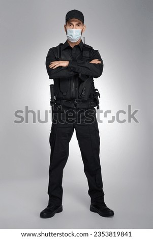 Strong young male cop wearing protective mask looking at camera in studio. Enforcement police officer in uniform standing with crossed hands, on gray background. Concept of work, quarantine.