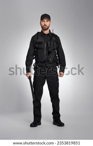 Confident law enforcement officer in black uniform standing with baton in studio. Front view of bearded policeman holding truncheon and looking at camera, on gray background. Police equipment concept.