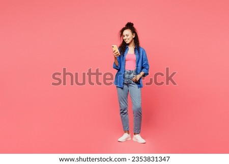 Full body young smiling happy woman of African American ethnicity wear blue shirt casual clothes hold in hand use mobile cell phone isolated on plain pastel pink background studio. Lifestyle concept