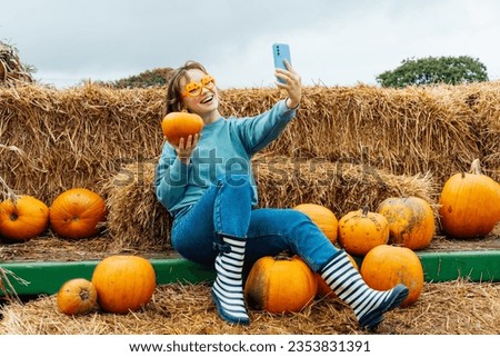 Smiling woman in fun glasses sitting on straw bales and holding pumpkin and making selfie on phone. Selecting Thanksgiving and Halloween holidays decor on agriculture farm. Autumn fall festive mood