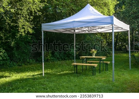 White gazebo or pavilion with a wooden table, benches and a flower bouquet on the meadow in a natural garden, idyllic summer place gives protection from sun and rain, copy space, selected focus Royalty-Free Stock Photo #2353839713