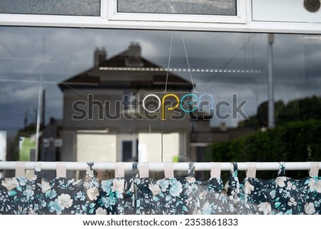 "open" sign neon multicolored lights white yellow teal green turquoise and blue hanging from inside of shop salon office window curtain rod blue floral blinds shades home reflected in glass shop front