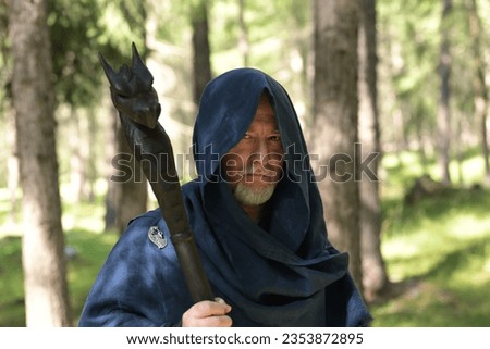 Half-bust portrait of a middle-aged Caucasian man wearing a blue hooded cloak and holding a black wizard staff in one hand, he stands in a wood.
Space for copy. Royalty-Free Stock Photo #2353872895