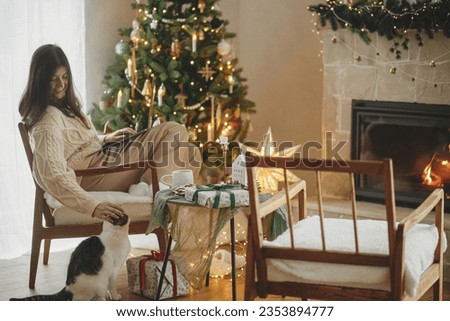 Happy woman working on laptop and caressing cute little cat in festive decorated christmas room with tree lights. Christmas holidays and pet. Remote work in winter holidays