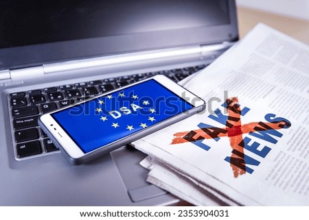 Smartphone with DSA and EU flag on screen over a newspaper with Fake news and a laptop. Fake news, HOAX concept Royalty-Free Stock Photo #2353904031