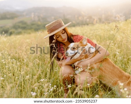 Beautiful girl with long black hair wearing in red dress and a hat.  Corgi dog in the background of green grass. A girl gently strokes her corgi dog. Walking the dog, friendship. Summer picture