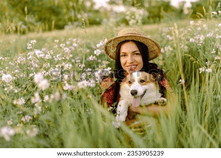 Beautiful girl with long black hair wearing in red dress and a hat.  Corgi dog in the background of green grass. A girl gently strokes her corgi dog. Walking the dog, friendship. Summer picture