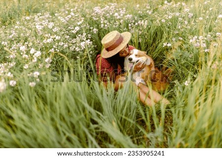 Beautiful girl with long black hair wearing in red dress and a hat.  Corgi dog in the background of green grass. A girl is kissing her corgi dog.  Walking the dog, friendship. Summer picture