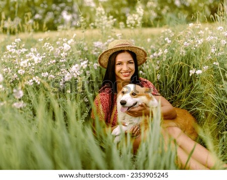 Beautiful girl with long black hair wearing in red dress and a hat.  Corgi dog in the background of green grass. A girl is holding her corgi dog.  Walking the dog, friendship. Summer picture