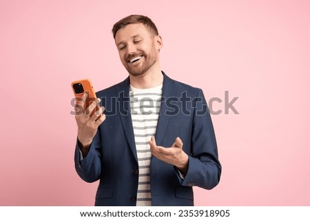 Joyful smiling man looking at mobile phone screen laughing rejoicing at good offer approval of deal over isolated pink background. Delighted trader with smartphone business communicates via video link Royalty-Free Stock Photo #2353918905