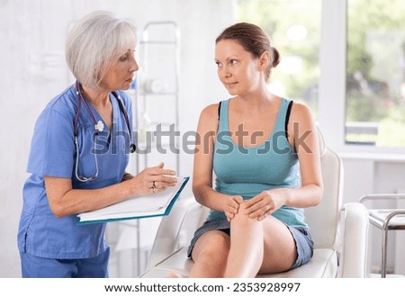 Mature female doctor conducts survey, listens to complaints of knee pain of female patient before procedure and fill in data in client medical record Royalty-Free Stock Photo #2353928997