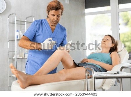 Concentrated experienced professional cosmetologist doing knee contouring injections to woman in aesthetic medicine office, injecting fat dissolving or lifting substances