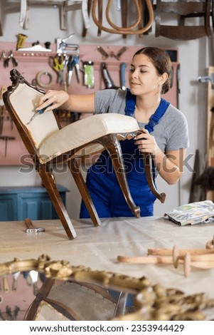 To replace upholstery, young female restorer carefully removes old cloth seat covering from antique wooden chair. Royalty-Free Stock Photo #2353944429