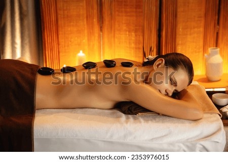 Hot stone massage at spa salon in luxury resort with warm candle light, blissful woman customer enjoying spa basalt stone massage glide over body with soothing warmth. Quiescent Royalty-Free Stock Photo #2353976015
