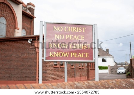 "NO CHRIST NO PEACE KNOW CHRIST KNOW PEACE" CHURCH SIGN OUTSIDE OF BRICK building with red background yellow gold text lettering behind wall red trim white house and car wires windows religious center Royalty-Free Stock Photo #2354072783