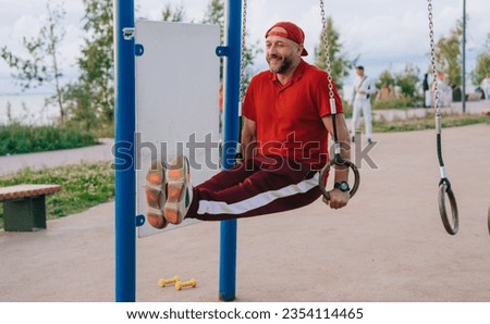 Cheerful mid aged caucasian man in sportswear training at sport court lifts legs leans on gymnastic rings at park. Strong bearded European guy at exercise outside. Healthy people, sport, CrossFit.