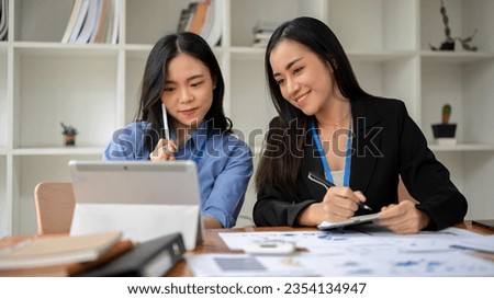 A pretty young Asian female office worker is working with her colleague, managing work on tablet and analyzing financial data on report in the office together.