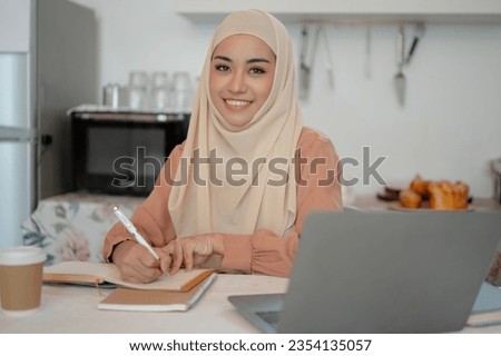 A beautiful Asian Muslim female college student is doing her homework at a table in the kitchen, writing something in her notebook.