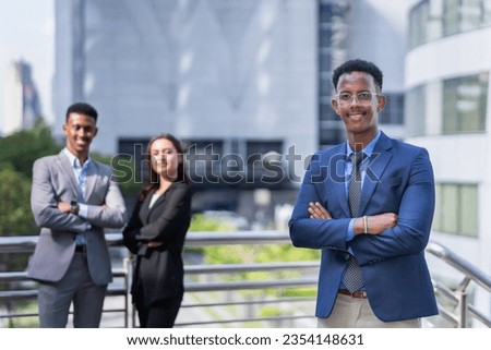 black executive man with glasses in blue suit smilng, standing, cross arm, looking at camera outside building in city. black businessman and caucasian businesswoman standing at background