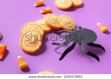 Tasty Halloween cookies with candies and paper spider on purple background