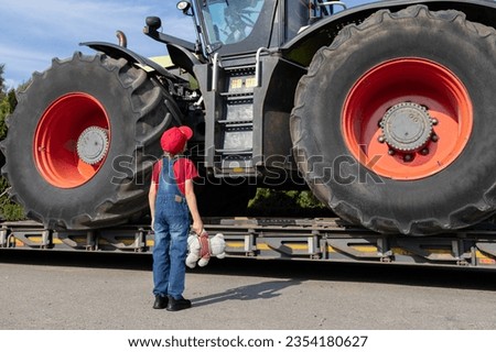 Child examines a huge tractor with large wheels loaded onto a truck for transportation. unrecognizable boy in denim overalls stands with his back in the frame. Royalty-Free Stock Photo #2354180627