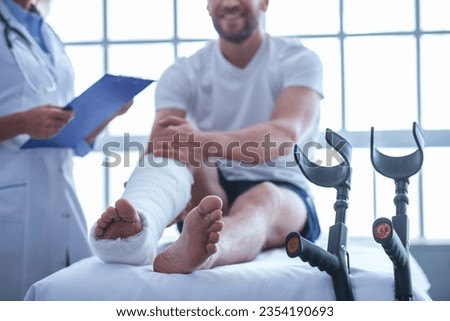 Cropped image of beautiful female medical doctor listening to handsome patient with broken leg and making notes while working in her office