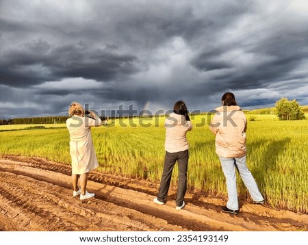 Tourist girls take pictures of rainbow and dramatic, gloomy clouds over a field on a spring, summer, autumn day. The concept of the beauty of nature and travel