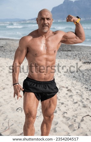 Attractive bald muscled fit guy in black rugby shorts doing a one-arm bicep flex on the beach.