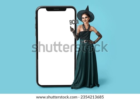 Beautiful young woman in Halloween costume of witch near big mobile phone on blue background