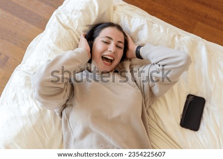 Young teen girl woman headphones listen to music at home lying in bedroom. Teenage girl relaxing meditating, looking at camera smiling Technology in daily life