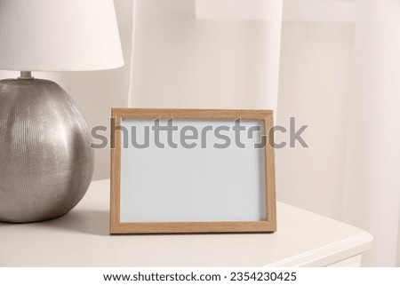 Empty square frame and lamp on white bedside table in room