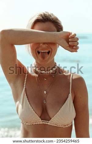Cheerful woman in bikini wearing golden necklace with palm trees and seashells on a sunlit beach.