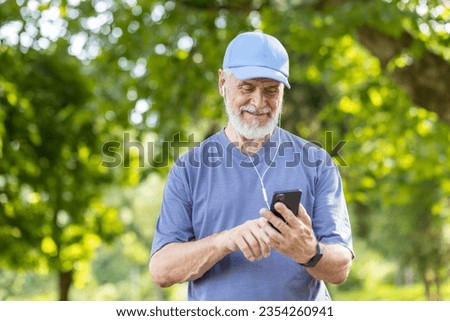 Senior gray-haired male athlete jogging in the park wearing headphones resting and listening to music online, using smartphone app, pensioner leading an active lifestyle.