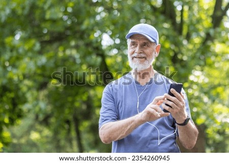 Senior gray-haired male athlete jogging in the park wearing headphones resting and listening to music online, using smartphone app, pensioner leading an active lifestyle.