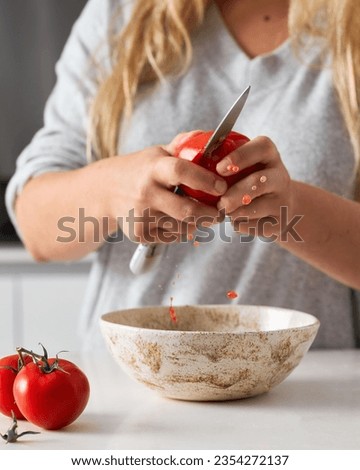 Kitchen scene of a caucasian woman cutting a tomato above a white textured ceramic bowl. Tomato juices leaking. White countertop. Tomatoes on the counter. Vertical still life. 