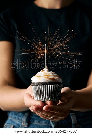 Festive image with a woman holding a chocolate cupcake topped with roasted meringue that has sparkler inside. Dark black background. Vertical image. Girl is wearing jeans and black shirt.