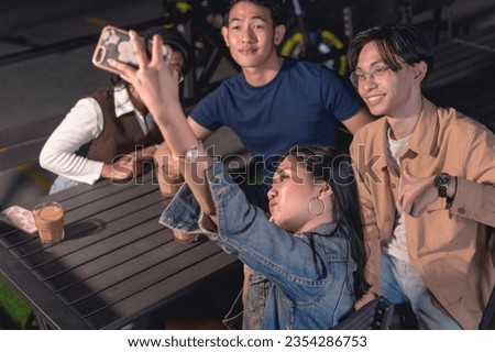 A couple of friends hanging out, two guys and two ladies. The lady in a denim jacket is holding up a phone to take a picture of them together. A rail, parking lot and motorcycle in the background.