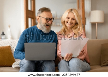 Happy couple of pensioners sitting using computer and holding papers at home interior, senior husband and wife with laptop reading email and checking bills, doing paperwork with enthusiasm together Royalty-Free Stock Photo #2354299677