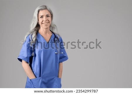 Healthcare and medicine concept. Happy middle aged nurse lady with hands in pockets, posing wearing blue workwear and standing on gray studio background, smiling to camera. Empty space