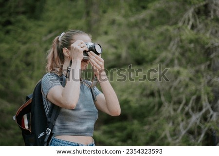 Blond girl photographs with an analog camera. Young hiker passionate about photography frames the scene. Woman with hair in a ponytail, striped t-shirt and backpack. Royalty-Free Stock Photo #2354323593
