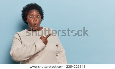 Studio shot of dark skinned African woman with curly hair points index finger on copy space reacts to something shocking dressed in casual sweatshirt isolated over blue background. Omg look at this